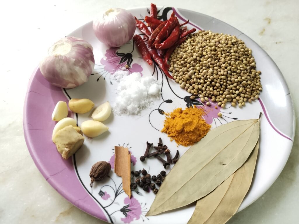 all spices for taro beef stew recipe on a plate including coriander seeds dried red chilies garlic cloves ginger onions turmeric salt bay leaves cinnamon black peppercorns cloves and black cardamom
